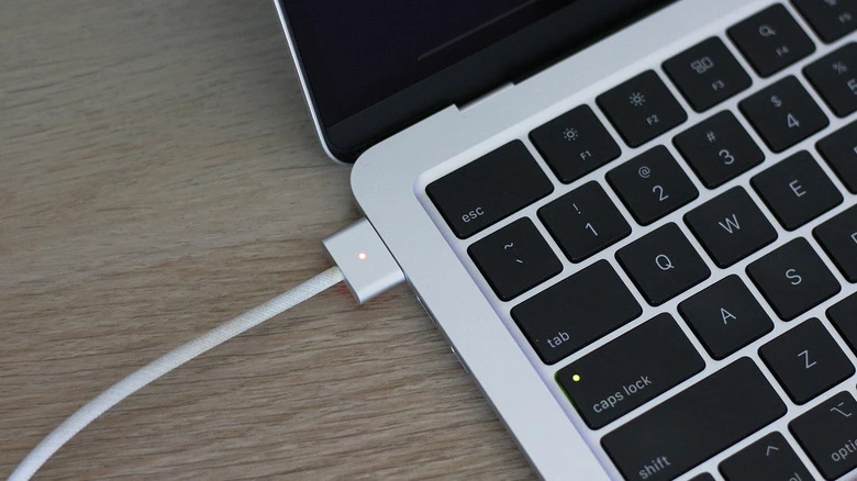 A white magnetic charging cable attached to the side of a silver laptop on a wooden desk